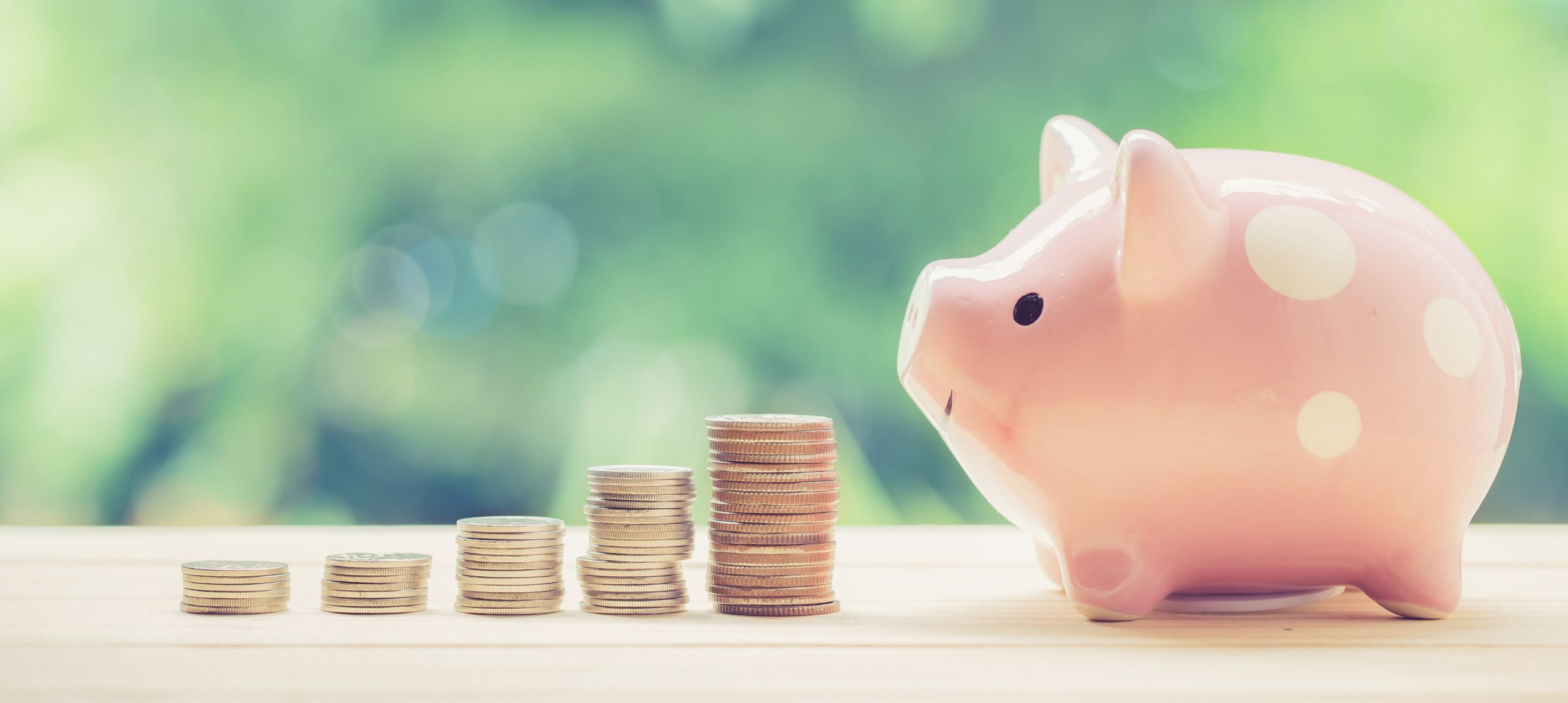 five stacks of coins growing towards the pink piggy bank sitting on the wooden table